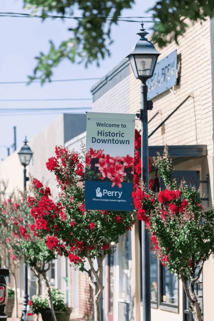 Historic downtown Perry, GA streetscape reflecting the local community Dinosaur Roofing & Repair proudly serves, featuring the Welcome to Historic Downtown Perry sign, red flowering trees, storefronts, and classic street lamps in Middle Georgia.